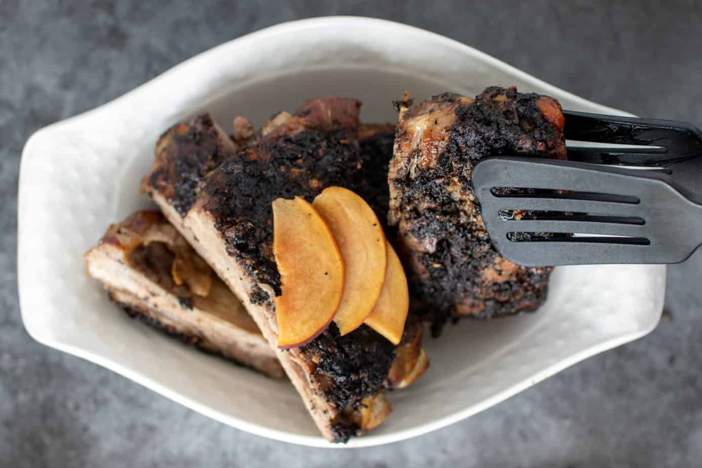 coffee rubbed rubs in a white dish- garnished with slices of peach, black spatula-tongs picking up one section of ribs