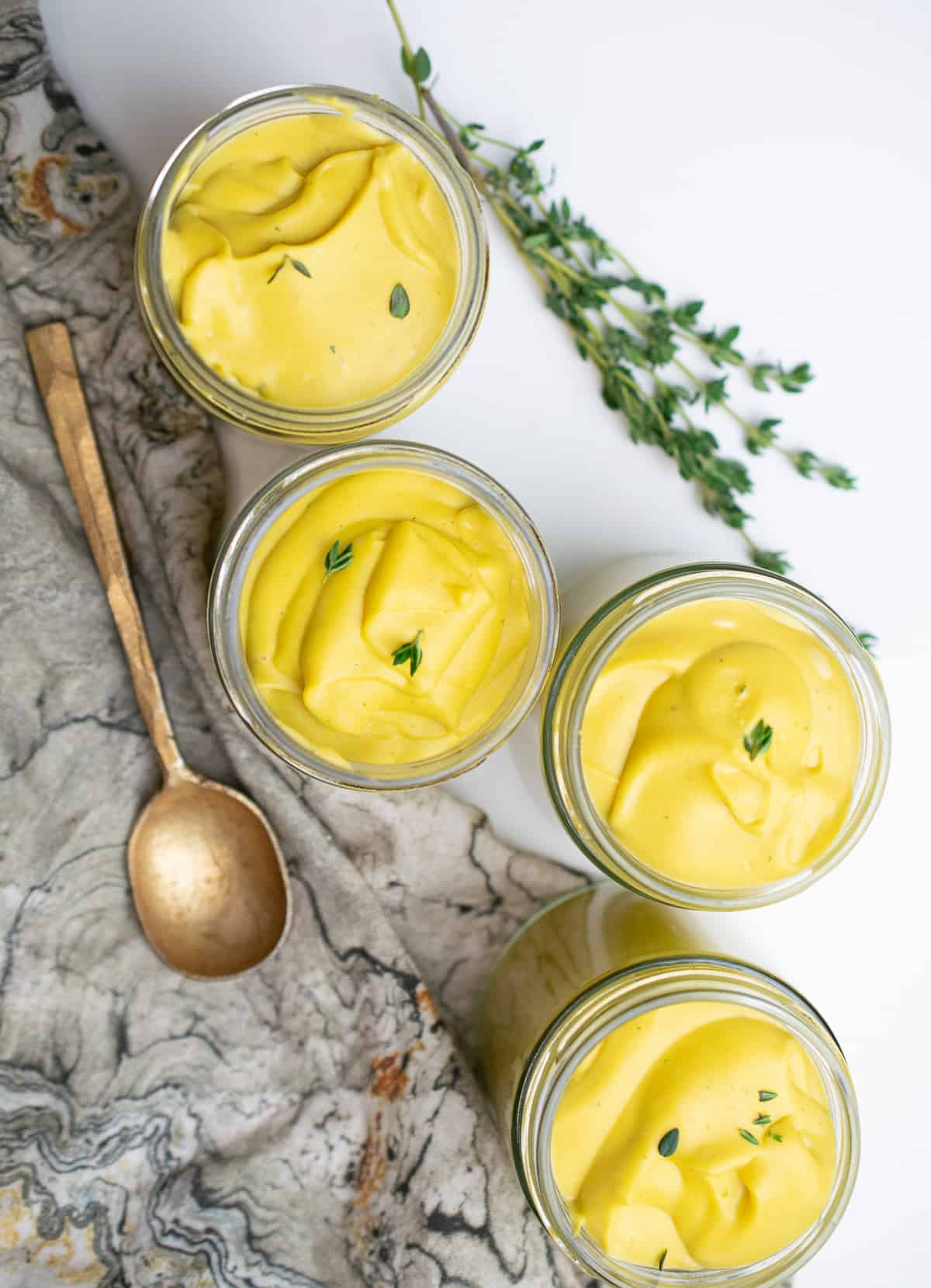 four jars of vegan butternut custard on a white counter with a napkin, golden spoon and sprigs of thyme