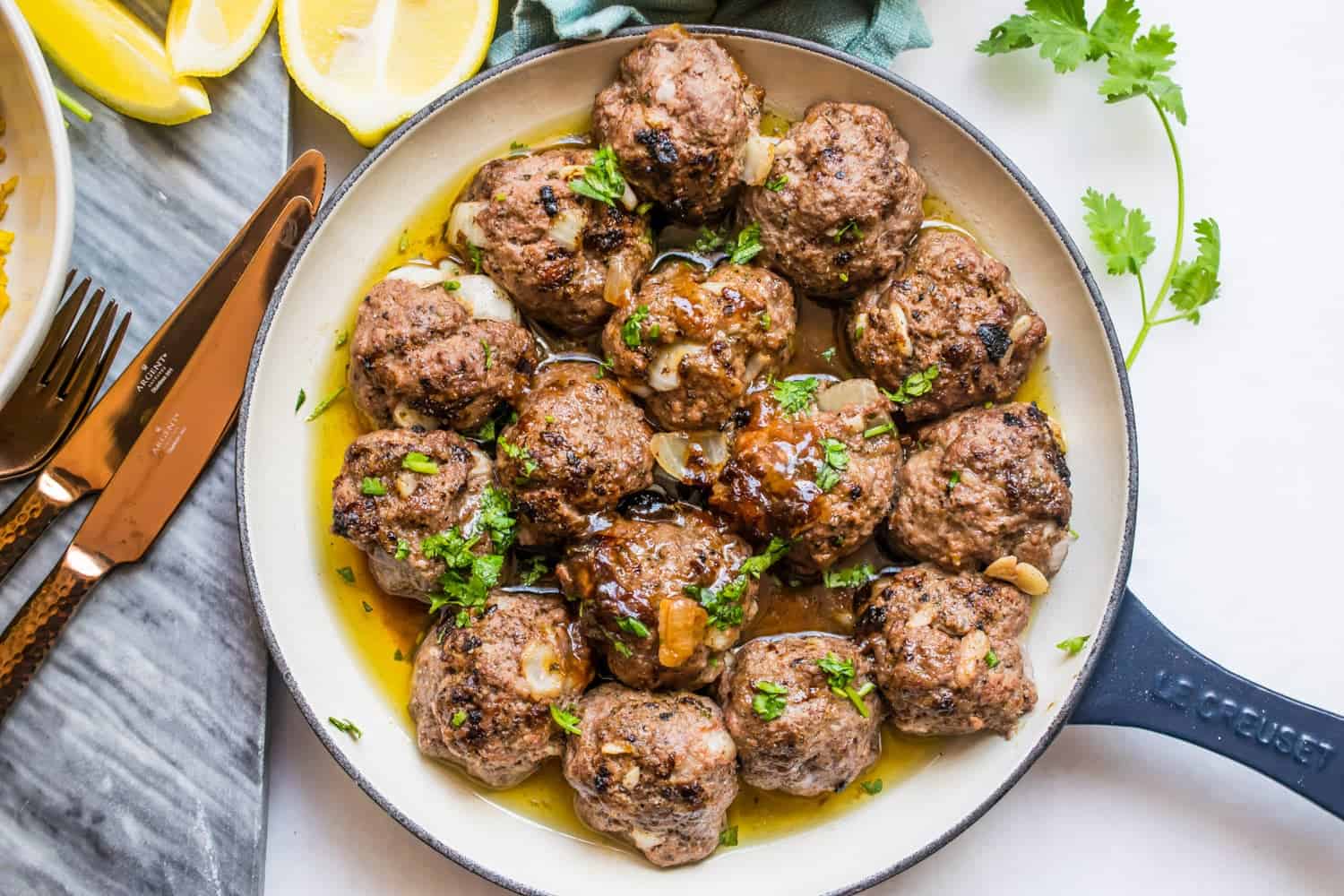 a blue le creuset pan holding 15 carne asada meatballs in their juice. garnished with onion and cilantro