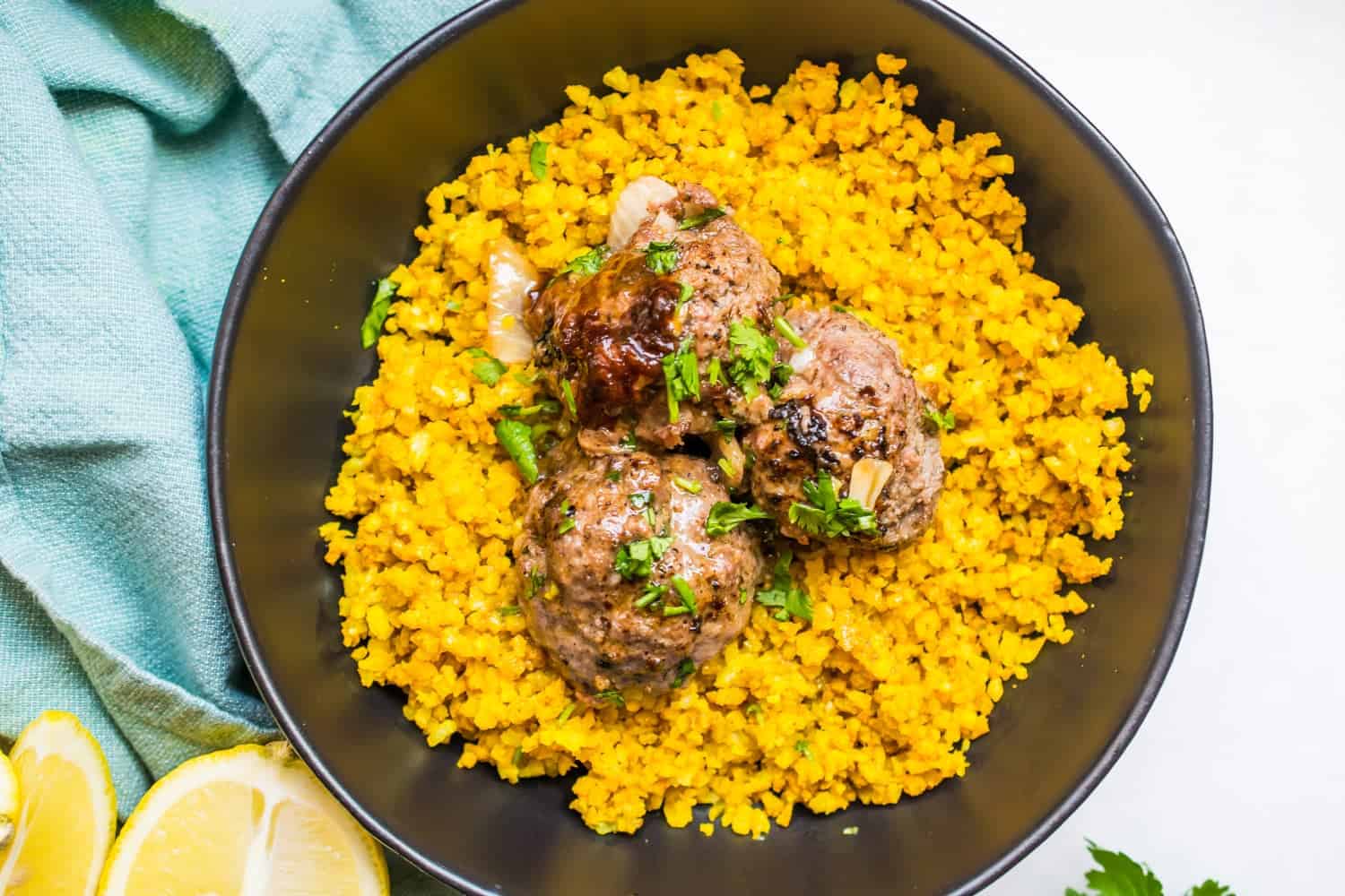 three carne asada meatballs over yellow rice garnished with cilantro in black bowl. half a lemon and blue cloth in background
