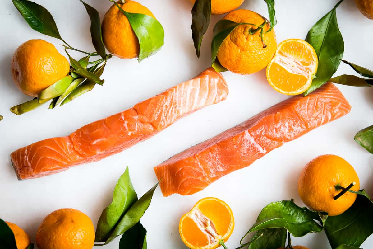 Overhead shot of beautifully coral wild caught salmon fillets on a white background, surrounded by fresh oranges with the leaves and stems intact.