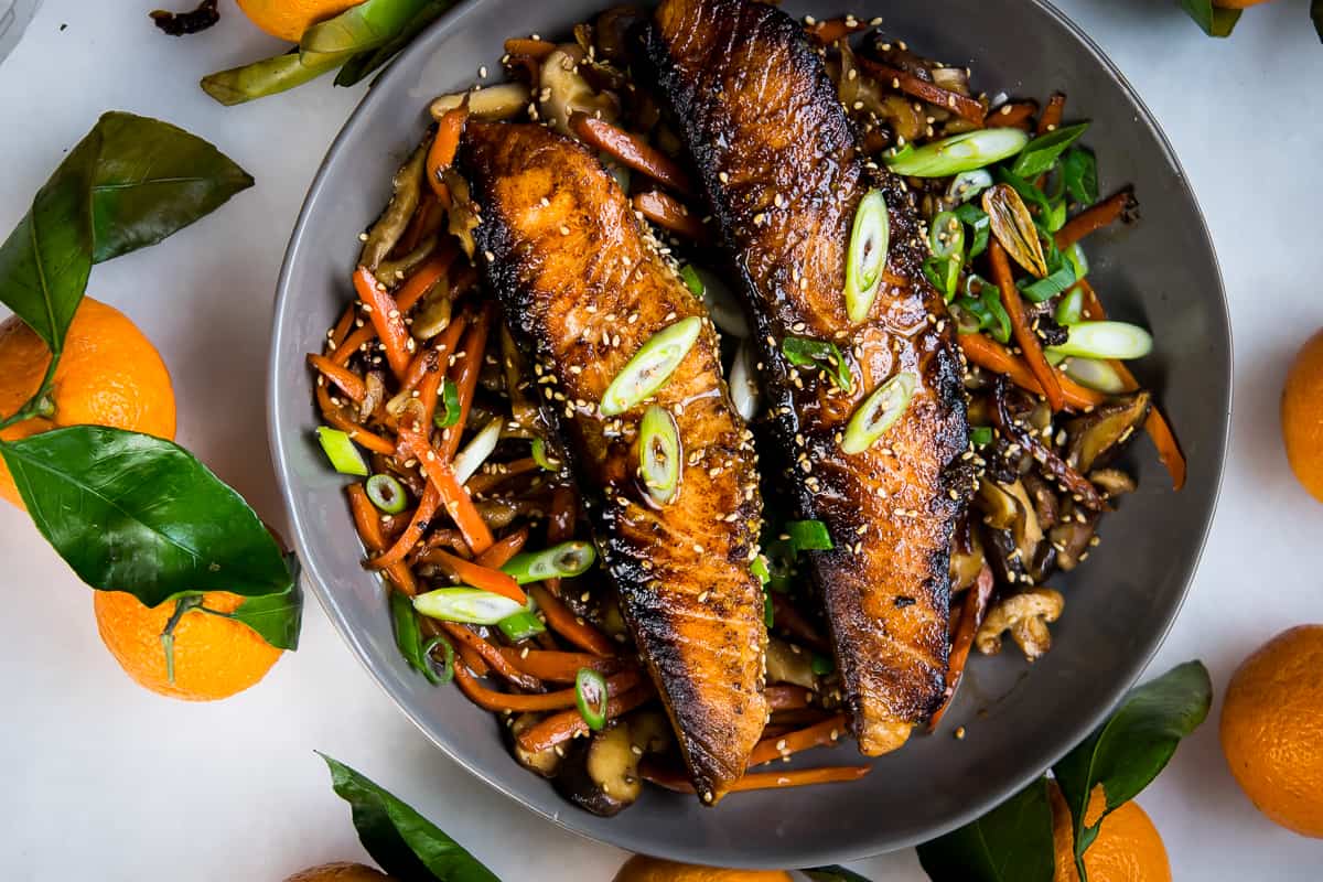 An overhead shot of Orange Ginger Salmon fillets, cooked till caramelized and almost blackened on the edges on a bed of veggies from the same recipe, garnished with green onions cut on a bias, served in a light charcoal colored bowl. The bowl sits on a white background, surrounded by fresh whole oranges with the steams and leaves intact.