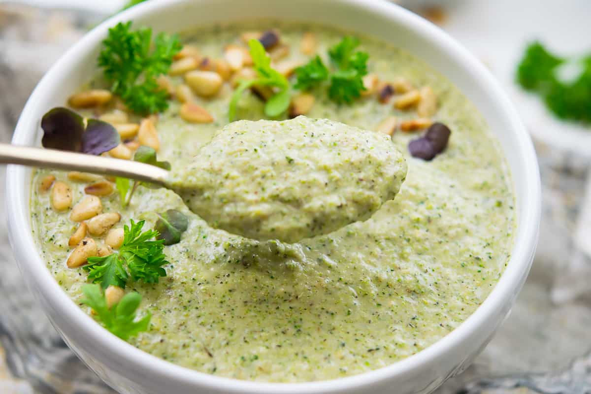 Overhead shot of a spoonful of creamy broccoli soup with a white bowl of soup, garnished with fresh herbs, microgreens, and toasted pine nuts. In the background, there's a gray napkin and fresh herbs out of focus.