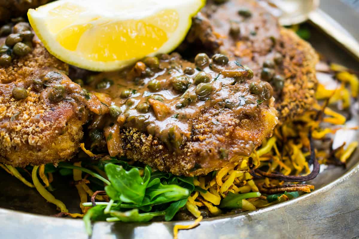 A silver plate with fried veggie noodles, topped with golden brown crispy paleo fried chicken breast, drizzled with silky caramel colored piccata sauce, bursting with capers, garnished with fresh herbs and a lemon wedge.