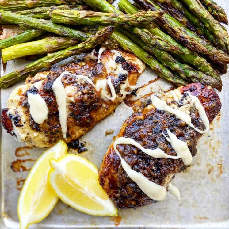 Macadamia Nut Garlic Chicken, seared to perfection, sits on an aluminum baking sheet lined with parchment paper. The chicken is drizzled with macadamia nut butter and garnished with lemon wedges, served alongside broiled asparagus.