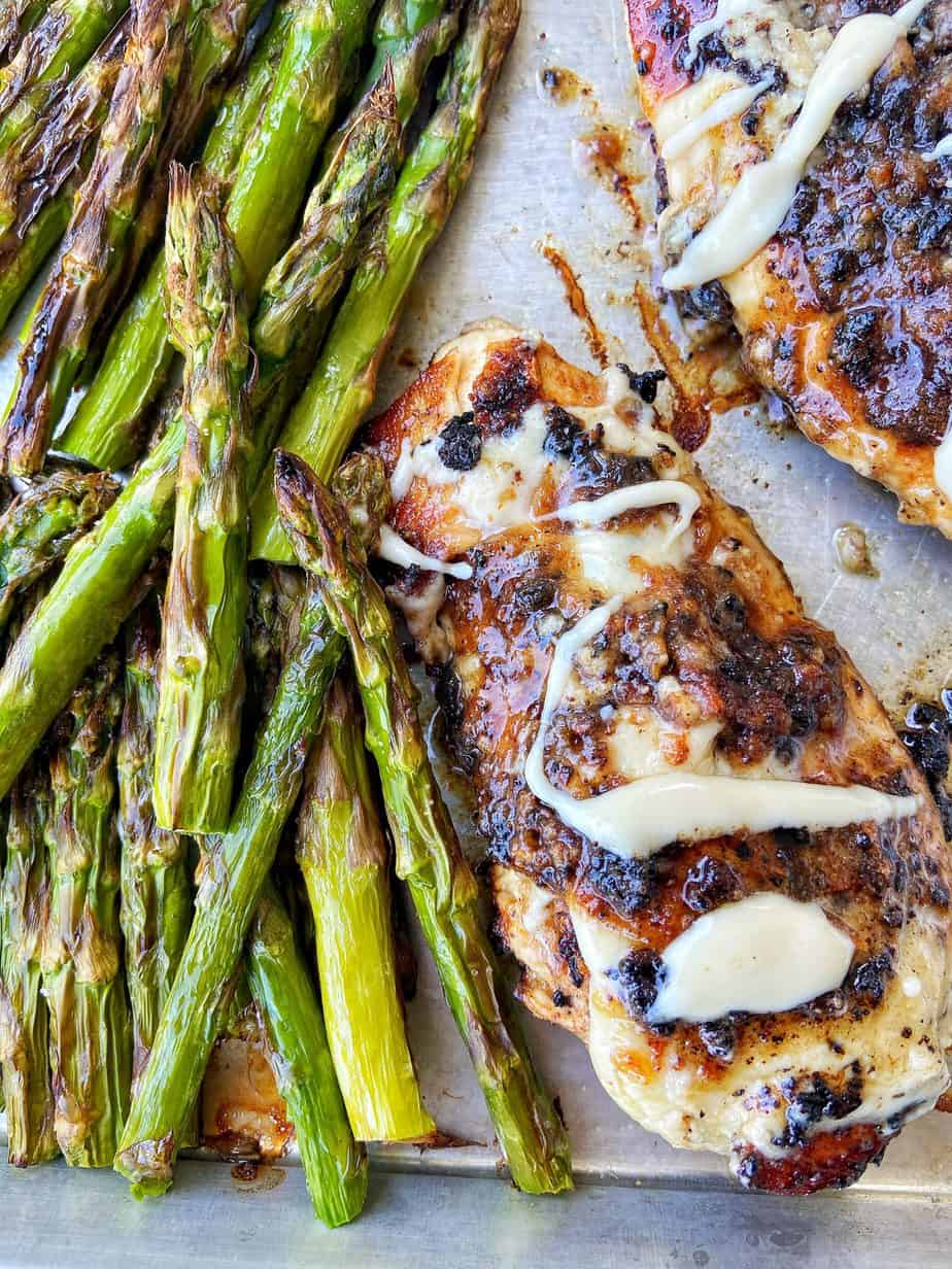 Macadamia Nut Garlic Chicken, seared to perfection, sits on an aluminum baking sheet lined with parchment paper. The chicken is drizzled with macadamia nut butter and served alongside broiled asparagus.