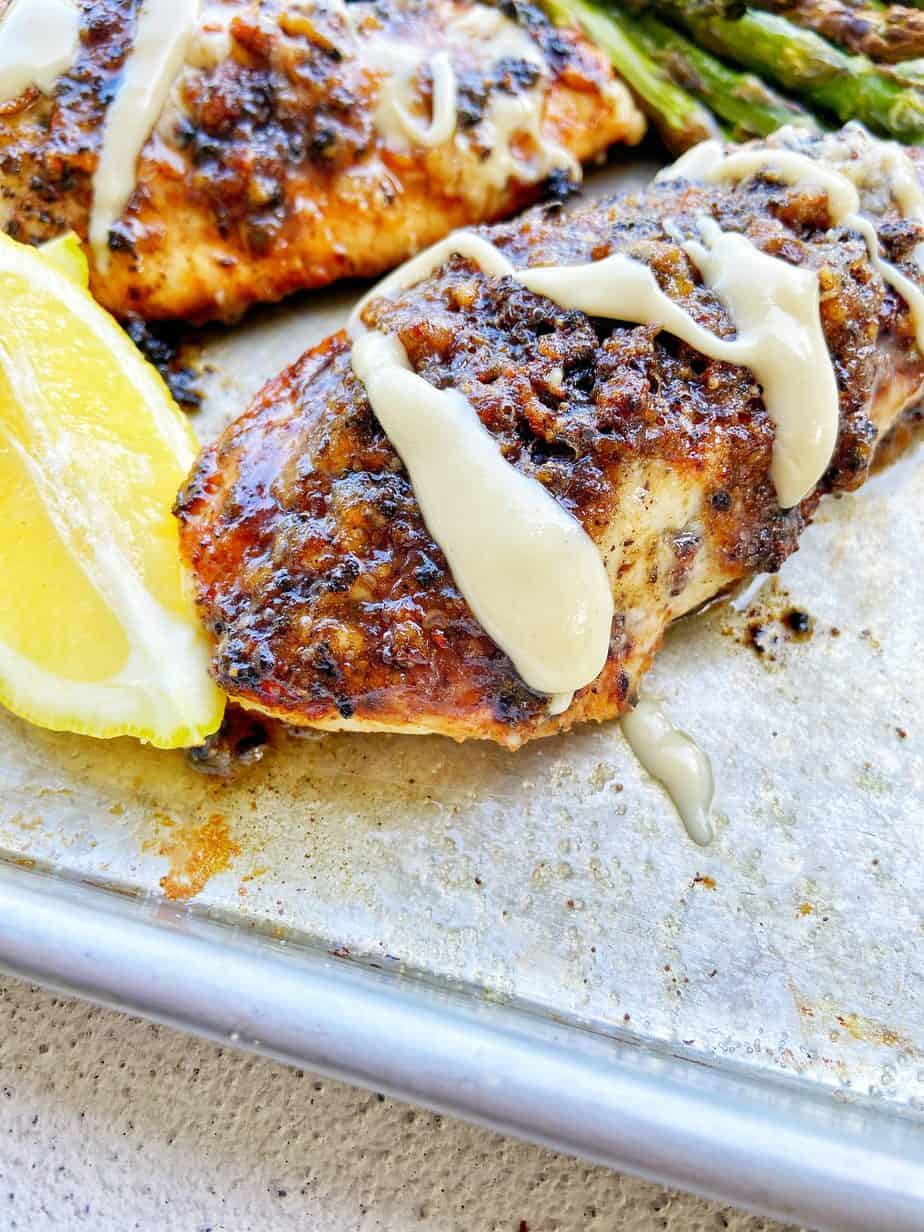 Macadamia Nut Garlic Chicken, seared to perfection, sits on an aluminum baking sheet lined with parchment paper. The chicken is drizzled with macadamia nut butter and garnished with lemon wedges and broiled asparagus sits in the background.