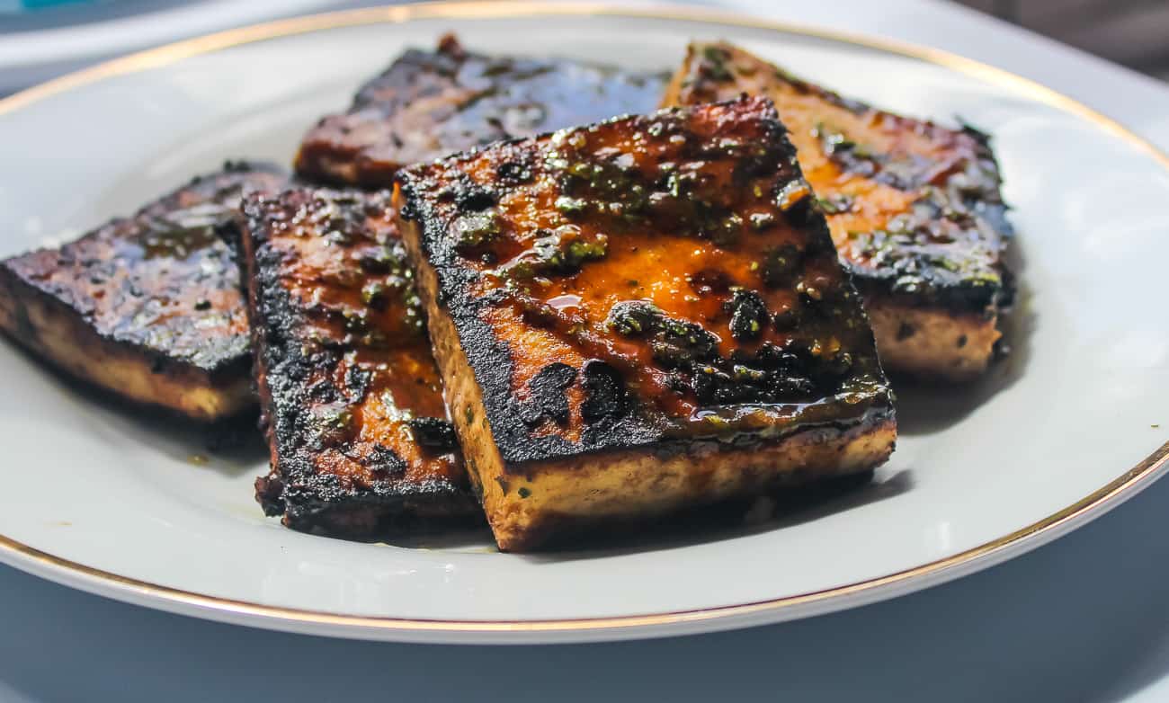 Several square blocks of pan fried glazed tofu sit on a white plate. The tofu is blackened and caramelized in some areas, with herbs and seasonings.