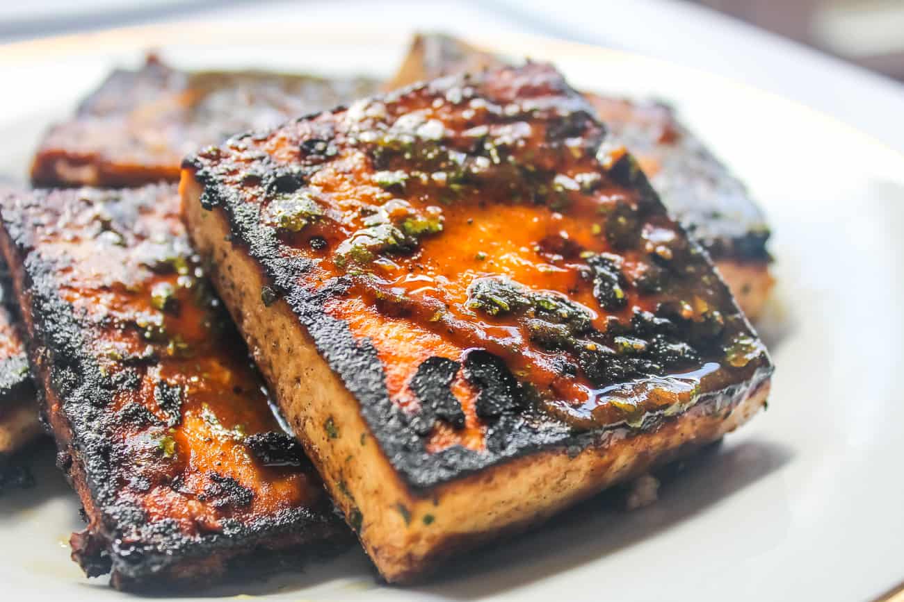 Several square blocks of pan fried glazed tofu sit on a white plate. The tofu is blackened and caramelized in some areas, with herbs and seasonings.