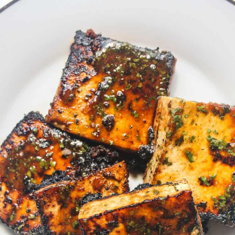 Overhead shot of several square blocks of pan fried glazed tofu sit on a white plate. The tofu is blackened and caramelized in some areas, with herbs and seasonings.