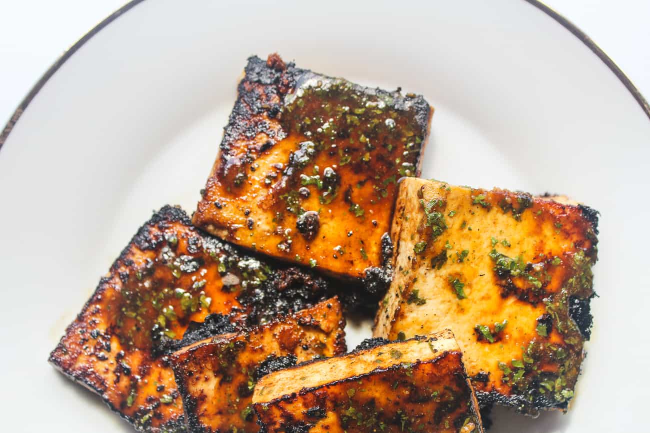 Overhead shot of several square blocks of pan fried glazed tofu sit on a white plate. The tofu is blackened and caramelized in some areas, with herbs and seasonings.