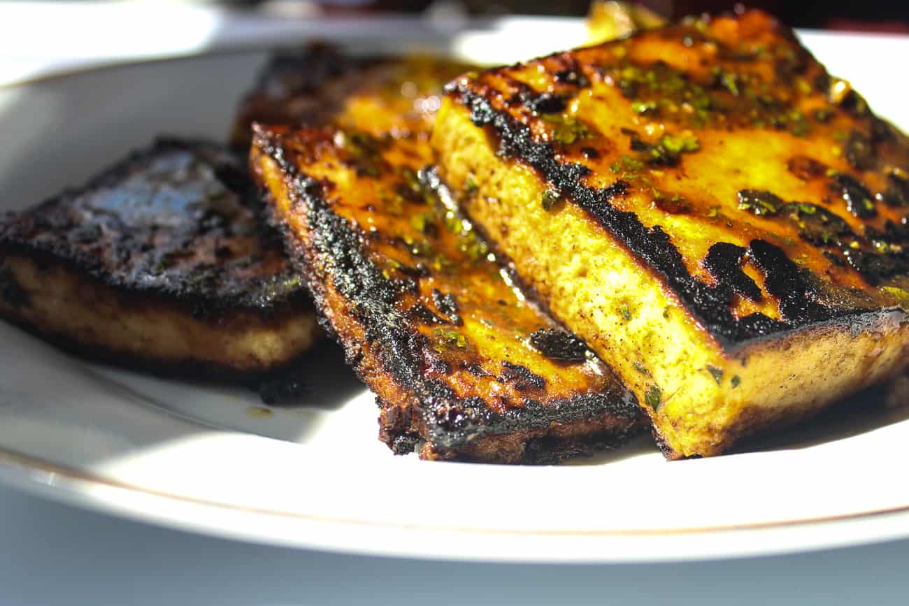 Several square blocks of pan fried glazed tofu sit on a white plate. The tofu is blackened and caramelized in some areas, with herbs and seasonings.