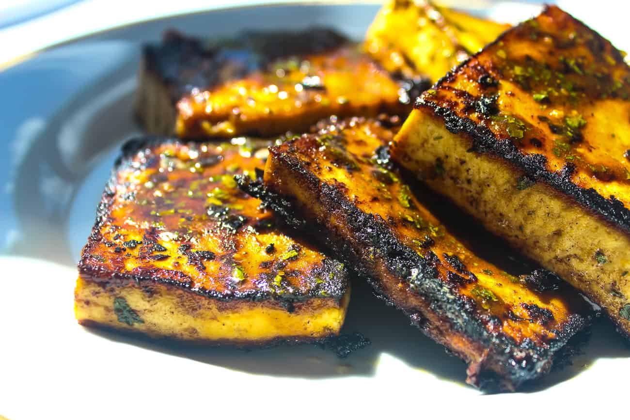 Several square blocks of pan fried glazed tofu sit on a white plate. The tofu is blackened and caramelized in some areas, with herbs and seasonings.