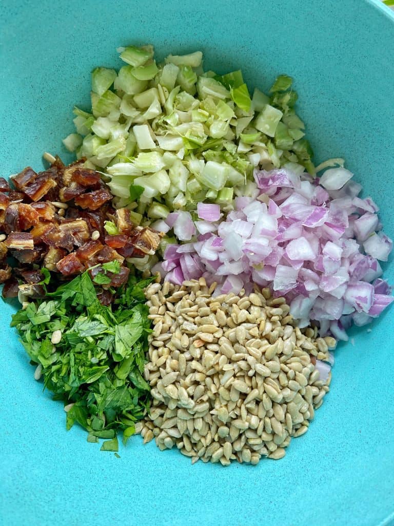 Ingredients for shredded chicken salad sit in a teal bowl: finely diced celery and red onion, minced fresh parsley, chopped pitted dates, and raw sunflower seeds.