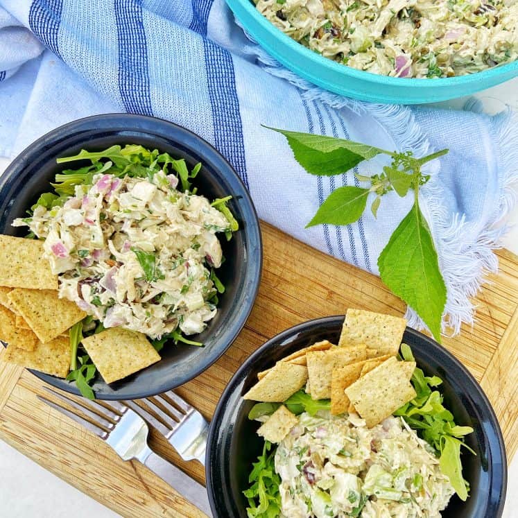 Two black bowls with a shredded chicken salad sitting on top of a bed of arugula, garnished with crackers. The bowls sit on a wooden cutting board with two forks, a gray and white striped cloth napkin, and a large bowl of shredded chicken salad to the side.