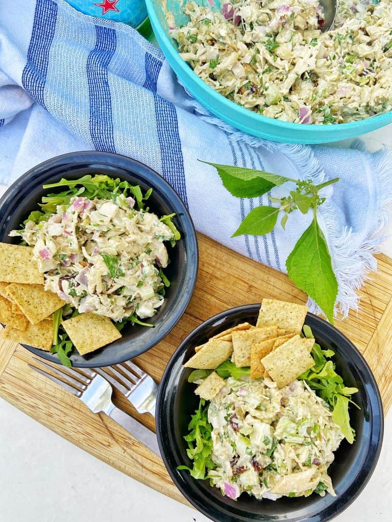 Two black bowls with a shredded chicken salad sitting on top of a bed of arugula, garnished with crackers. The bowls sit on a wooden cutting board with two forks, a gray and white striped cloth napkin, and a large bowl of shredded chicken salad to the side.