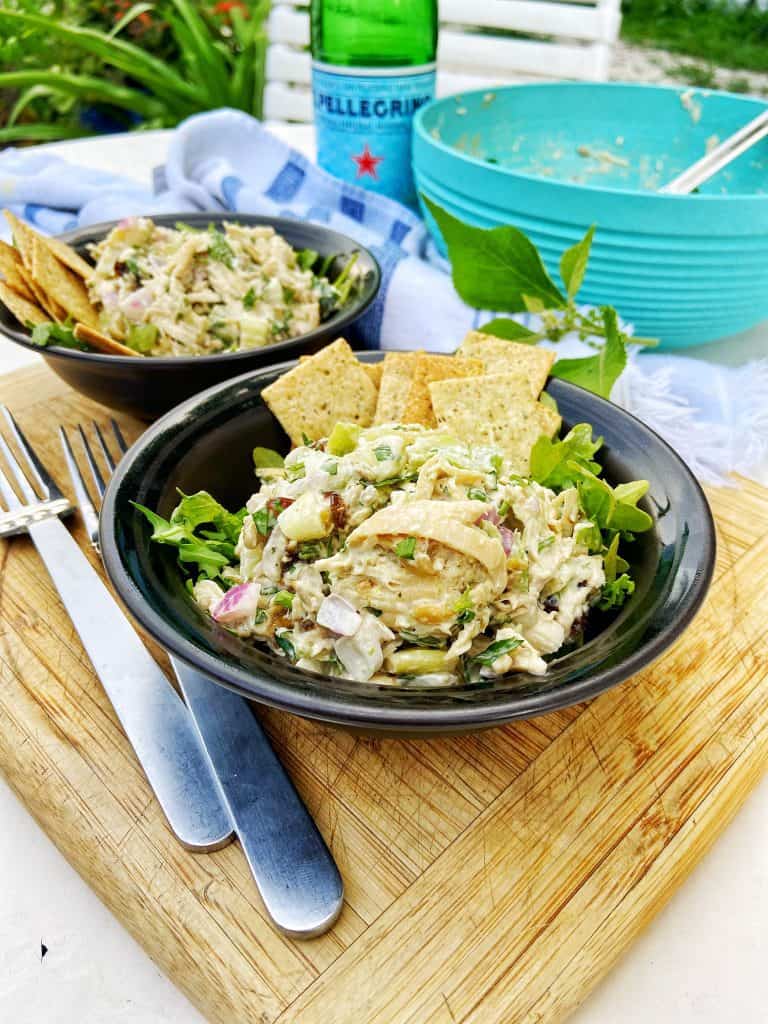 Two black bowls with a shredded chicken salad sitting on top of a bed of arugula, garnished with crackers. The bowls sit on a wooden cutting board with two forks, a gray and white striped cloth napkin, and a large bowl of shredded chicken salad in a large teal bowl on the side.