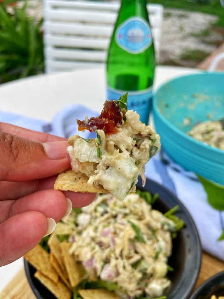 A cracker piled with shredded chicken salad. In the background is a lunch time scene: A black bowl with a shredded chicken salad sitting on top of a bed of arugula, garnished with crackers. In the background sits a gray and white striped cloth napkin, a bottle of Pellegrino mineral water, and a large bowl of shredded chicken salad to the side.