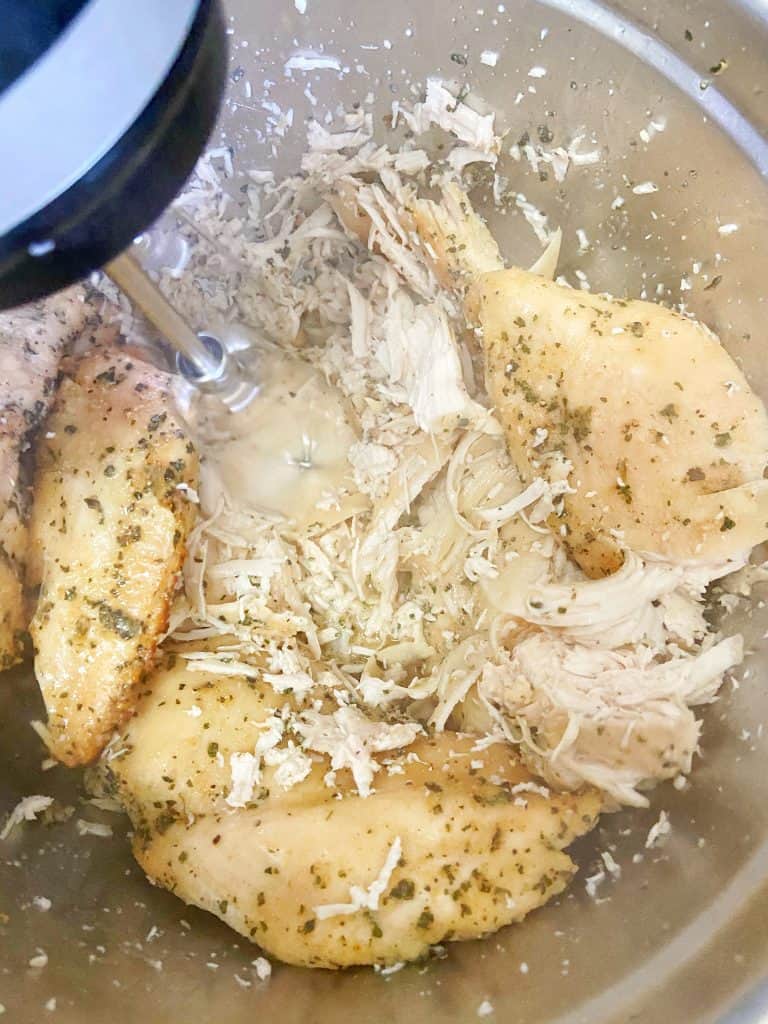 Freshly cooked (and steaming) seasoned chicken breasts sit in a large metal bowl. The chicken is being shredded with a hand mixer.