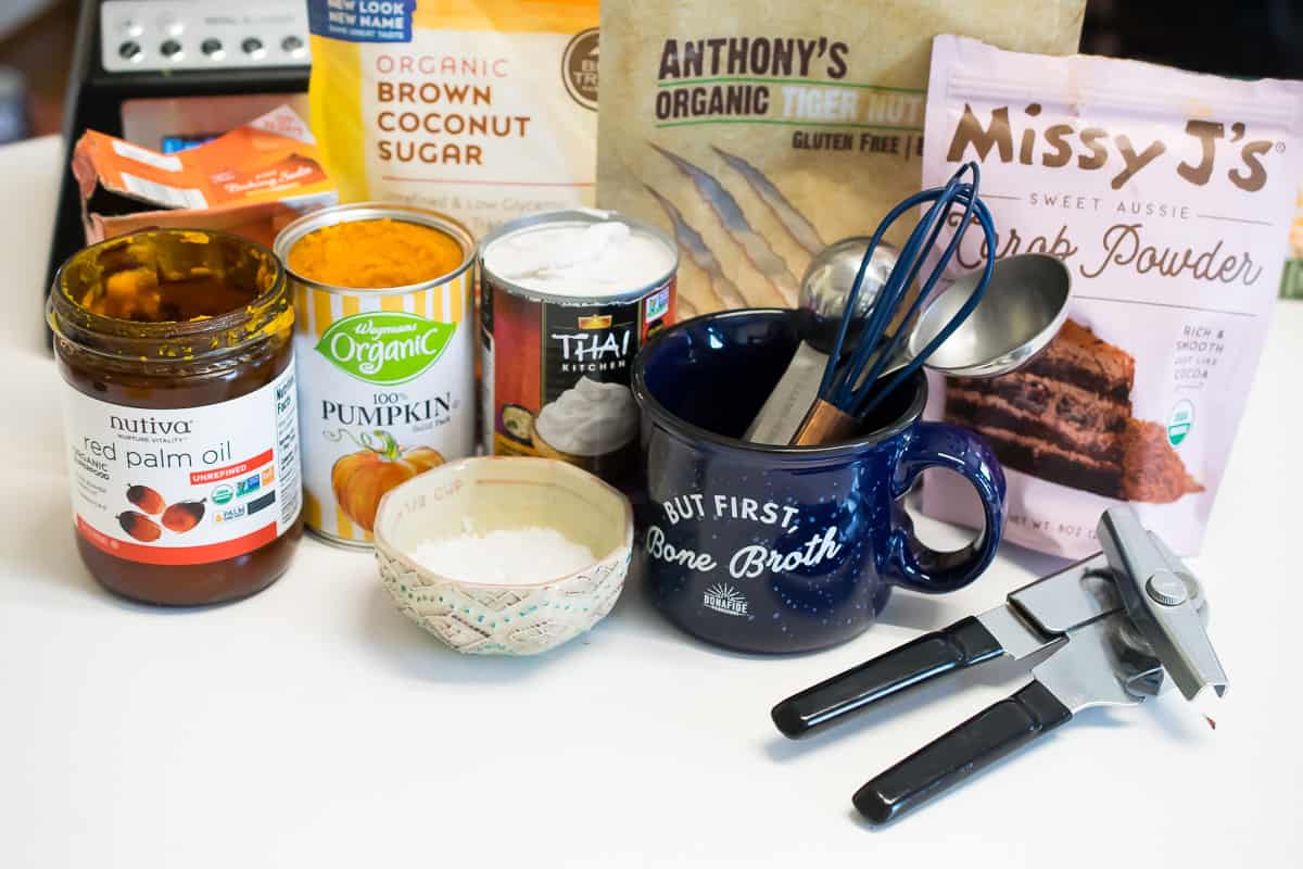 aip mug cake ingredients on table; red palm oil, canned pumpkin, coconut milk, salt, organic brown coconut sugar, tiger nut flour, carob flour, blue cup with measuring spoons, whisk, and can opener 