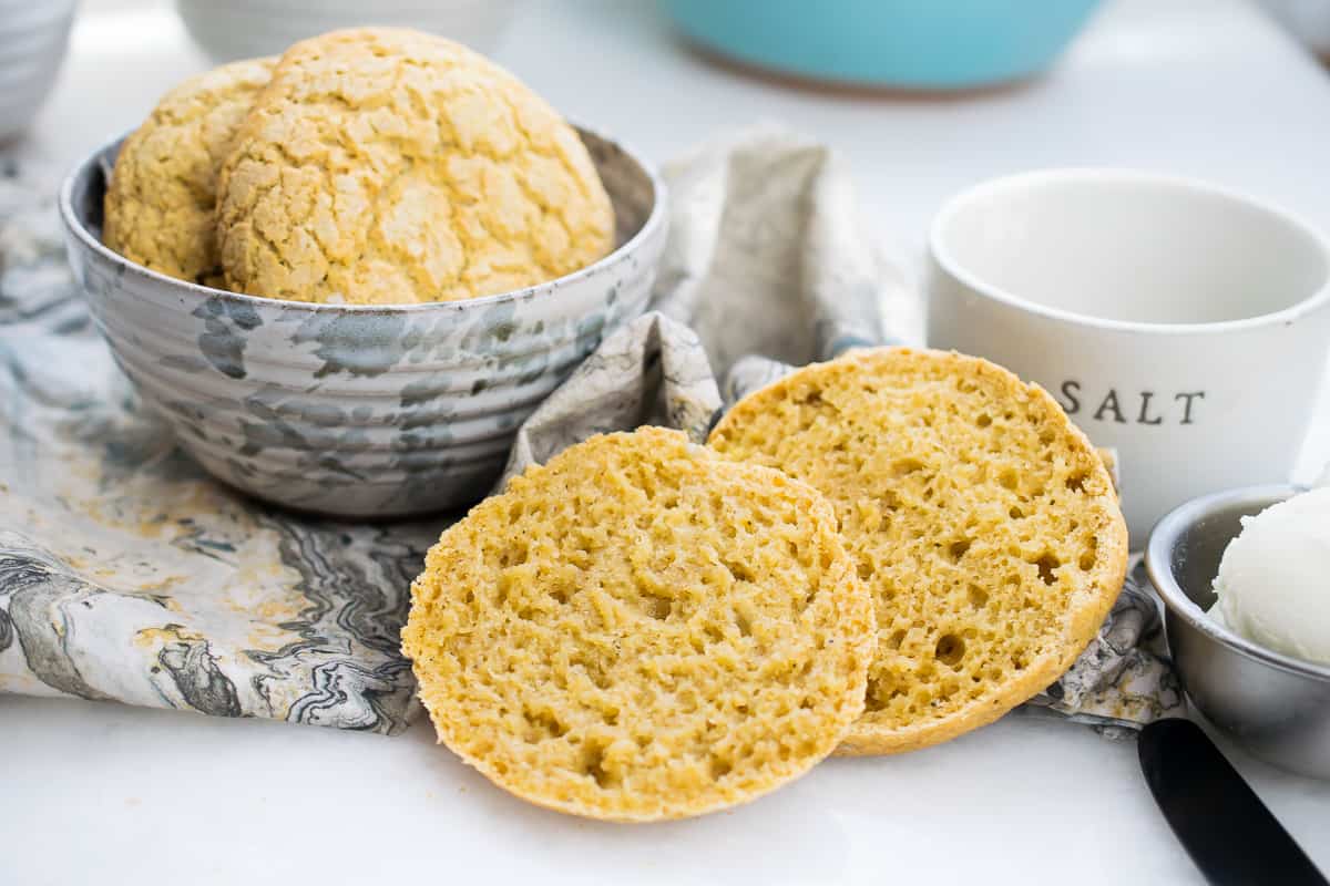 two AIP english muffins in small bowl, one muffin sliced in half, a tin with butter, and a tin with salt on side 