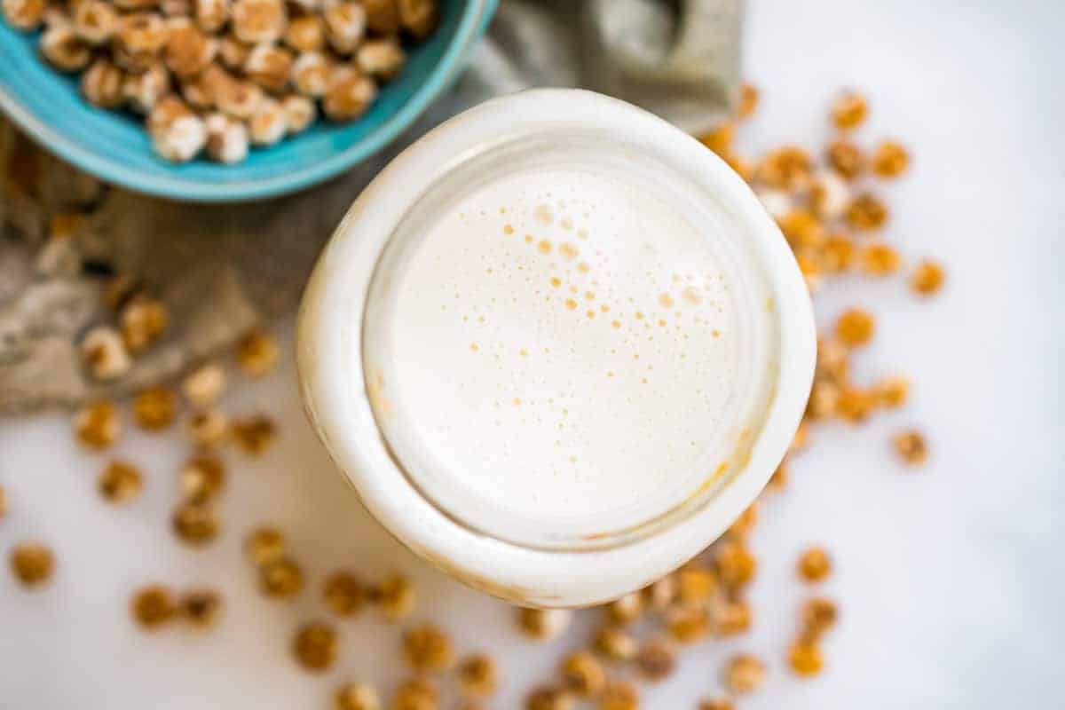 a shot of a full jar of tiger nut milk from above- frothy and white, in the background tiger nuts are on the table and in blue bowl