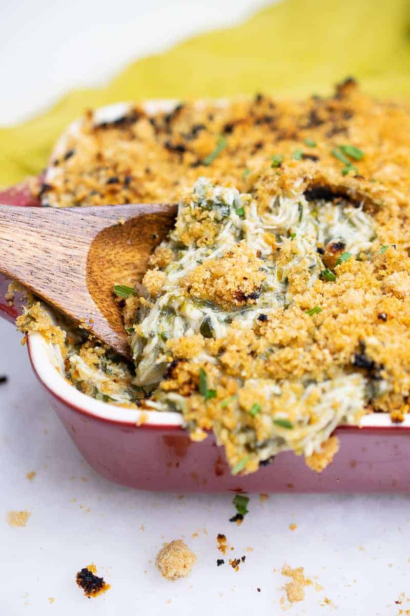 Scooping a wooden spoon into cheesy dairy-free AIP Chicken Casserole with spinach, baked and bubbly in a red casserole dish, topped with crunchy pork panko.