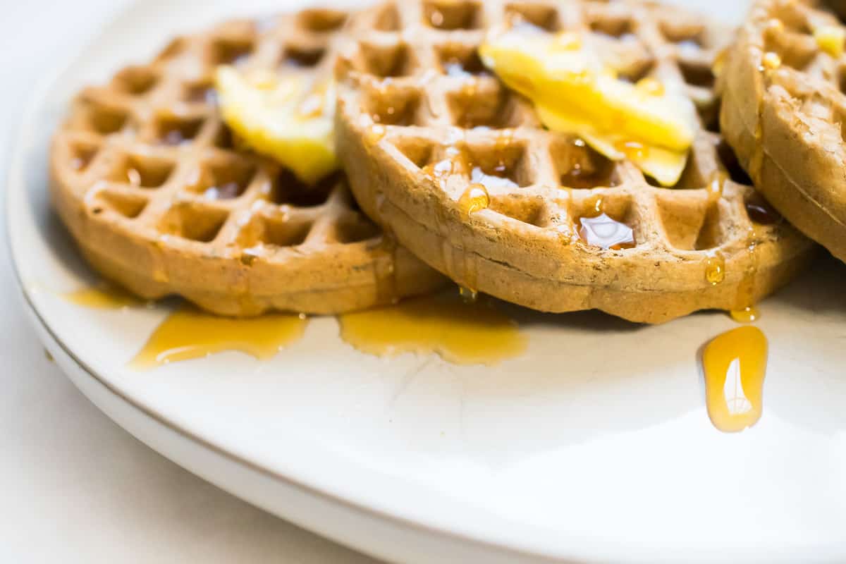 three nut free paleo waffles laying on each other topped with butter and maple syrup on a white plate