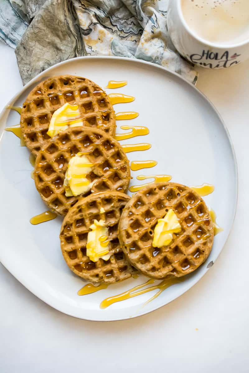 four nut free paleo waffles spread out on a white plate, each of them with a little butter on stop and drizzled with maple syrup