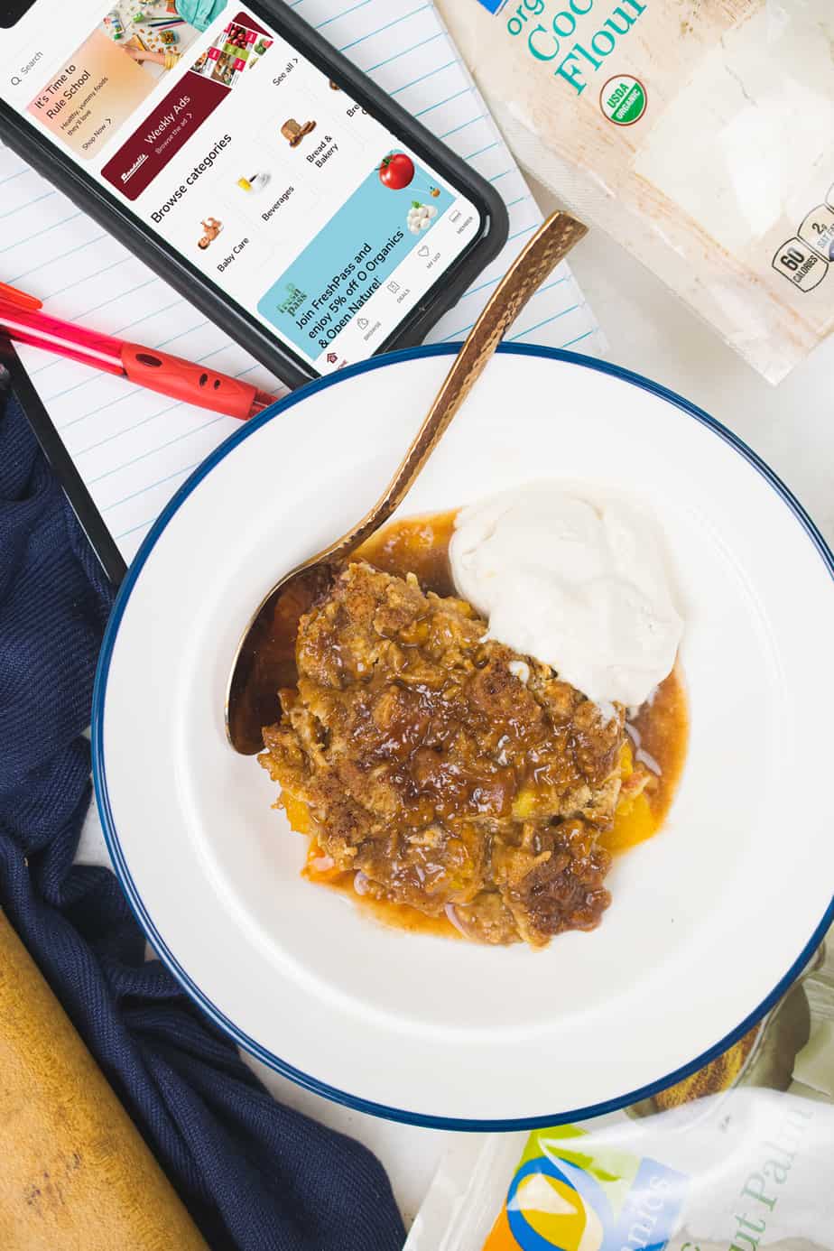 aip peach cobbler with icecream on the side in a bowl with a spoon set on a desk with a notepad a red ped and an iphone opened up to the Randalls app and a bag of coconut flour in background