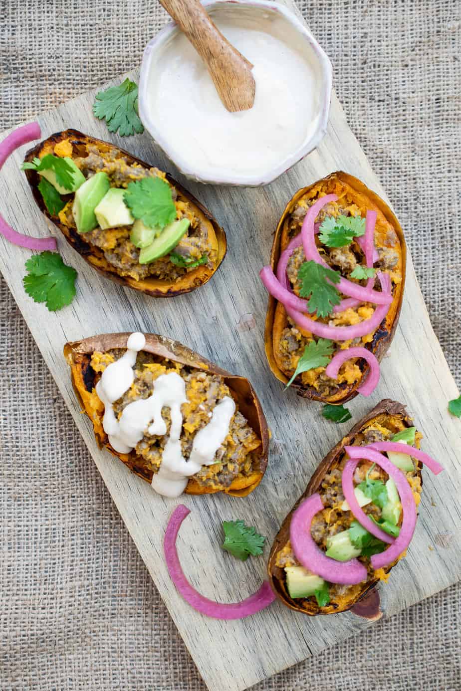 shot of final dish. four stuffed sweet potato skins. two garnished with cilantro and pickled red onion. one garnished with sauce. one garnished with cilantro and avocado. some cilantro and onion on the cutting board. cloth underneath cutting board. 