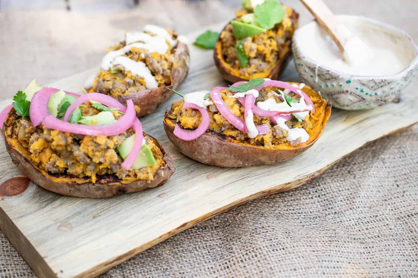 four stuffed sweet potato skins with ground sausage garnished with red onion, cilantro, and a mayo-based garnish. a bowl of the sauce on the side. all set on a plank of wood over cloth.
