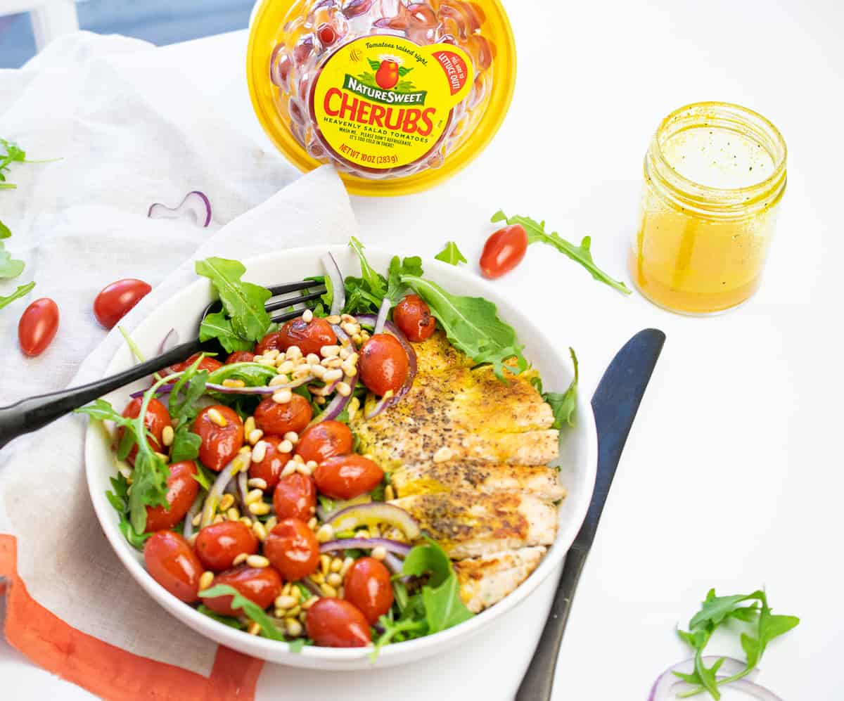 grilled chicken, cherry tomato and pine nut salad in a white bowl, with knife and fork and extra dressing on the side 