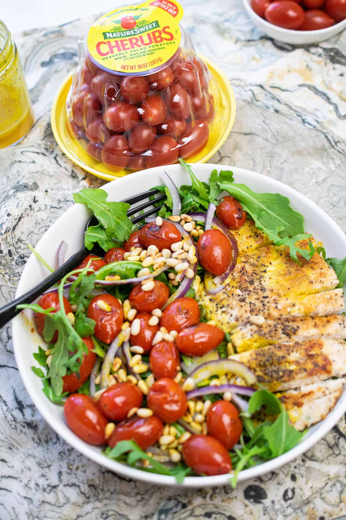 large bowl with grilled chicken, tomatoes, onion and pine nuts and cherub tomato container behind it