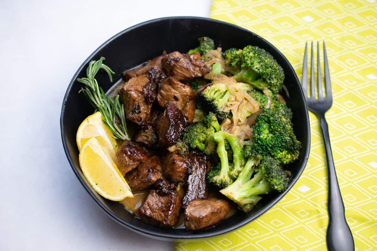 lemon herb beef and broccoli served in  a black bowl garnished with rosemary sprig and two lemon wedges.