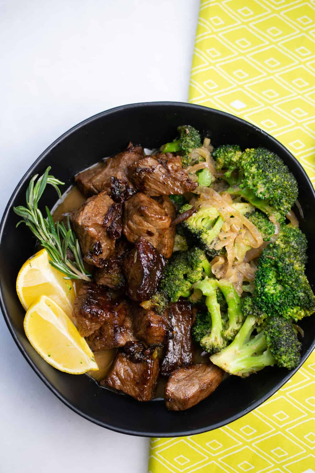 lemon herb beef and broccoli served in  a black bowl garnished with rosemary sprig and two lemon wedges.