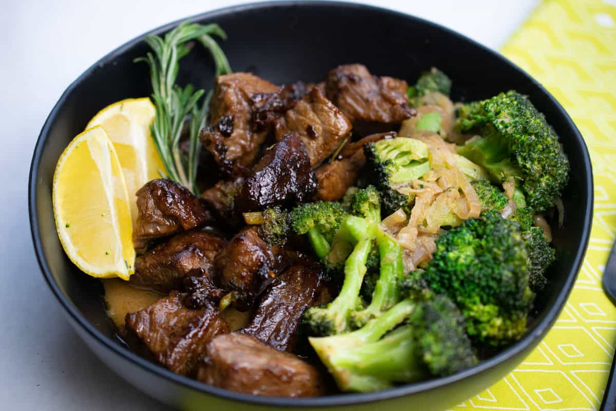Bowl of AIP Beef and Broccoli- beef in chunks and broccoli garnished with onion. Sprig of rosemary and lemon wedge on the side