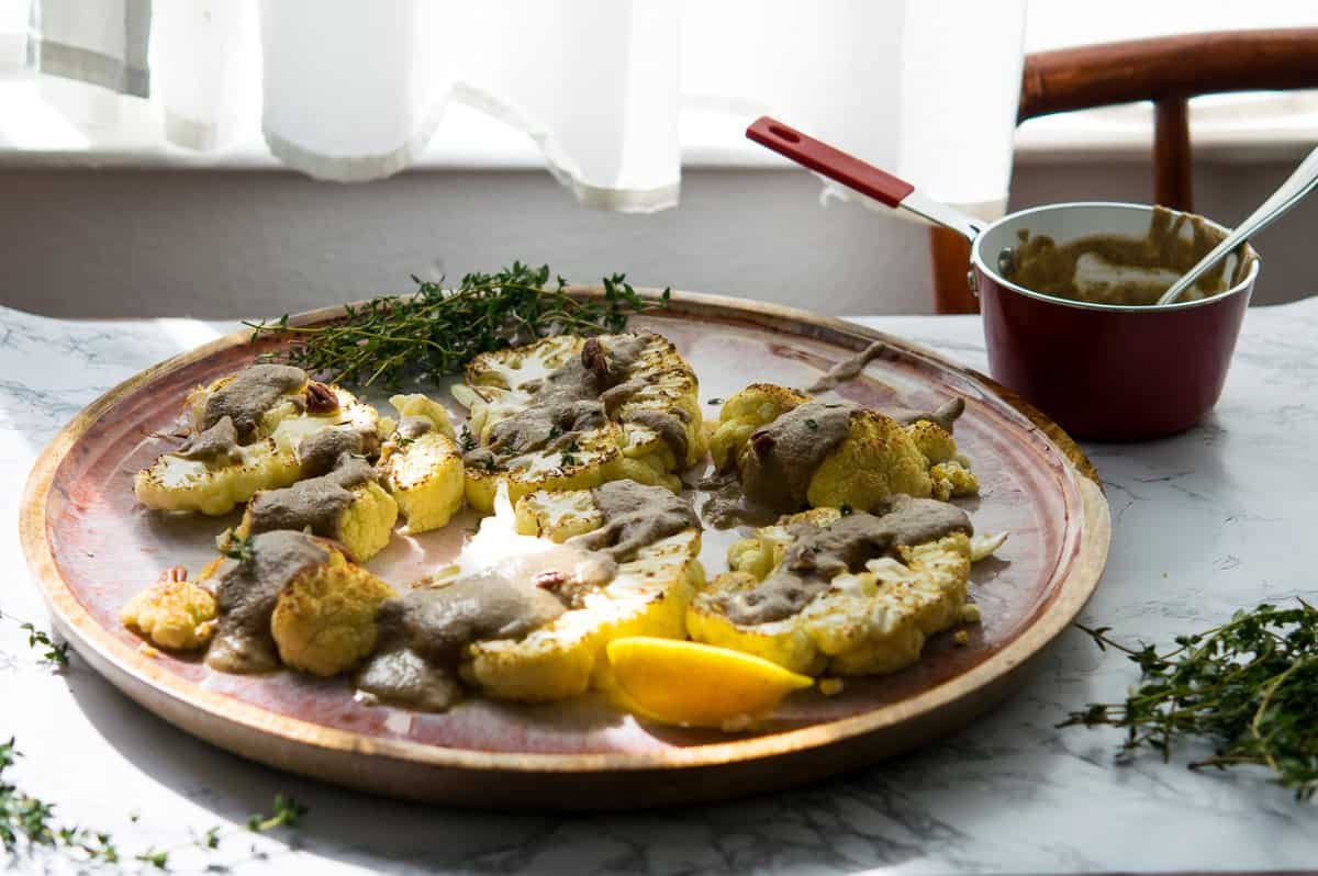 a table by a window with cauliflower steaks on a flatter with a thick drizzle of brown sauce and pecan pieces