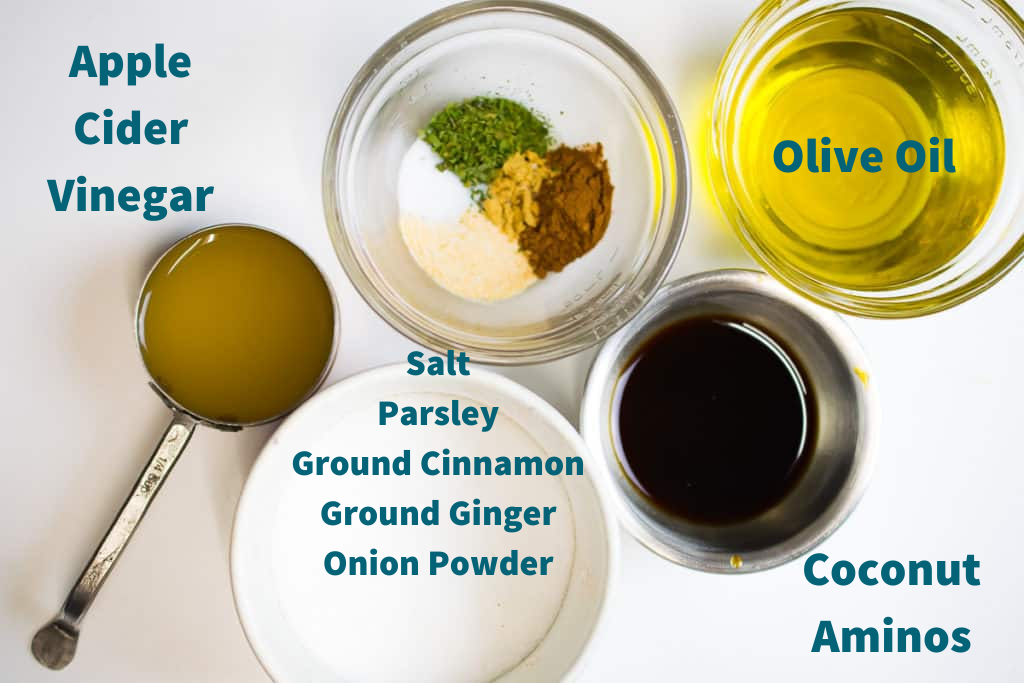 Ingredients for AIP salad dressing laid out on table; apple cider vinegar, olive oil, coconut aminos, salt, parsley, ground cinnamon, ground ginger, onion powder