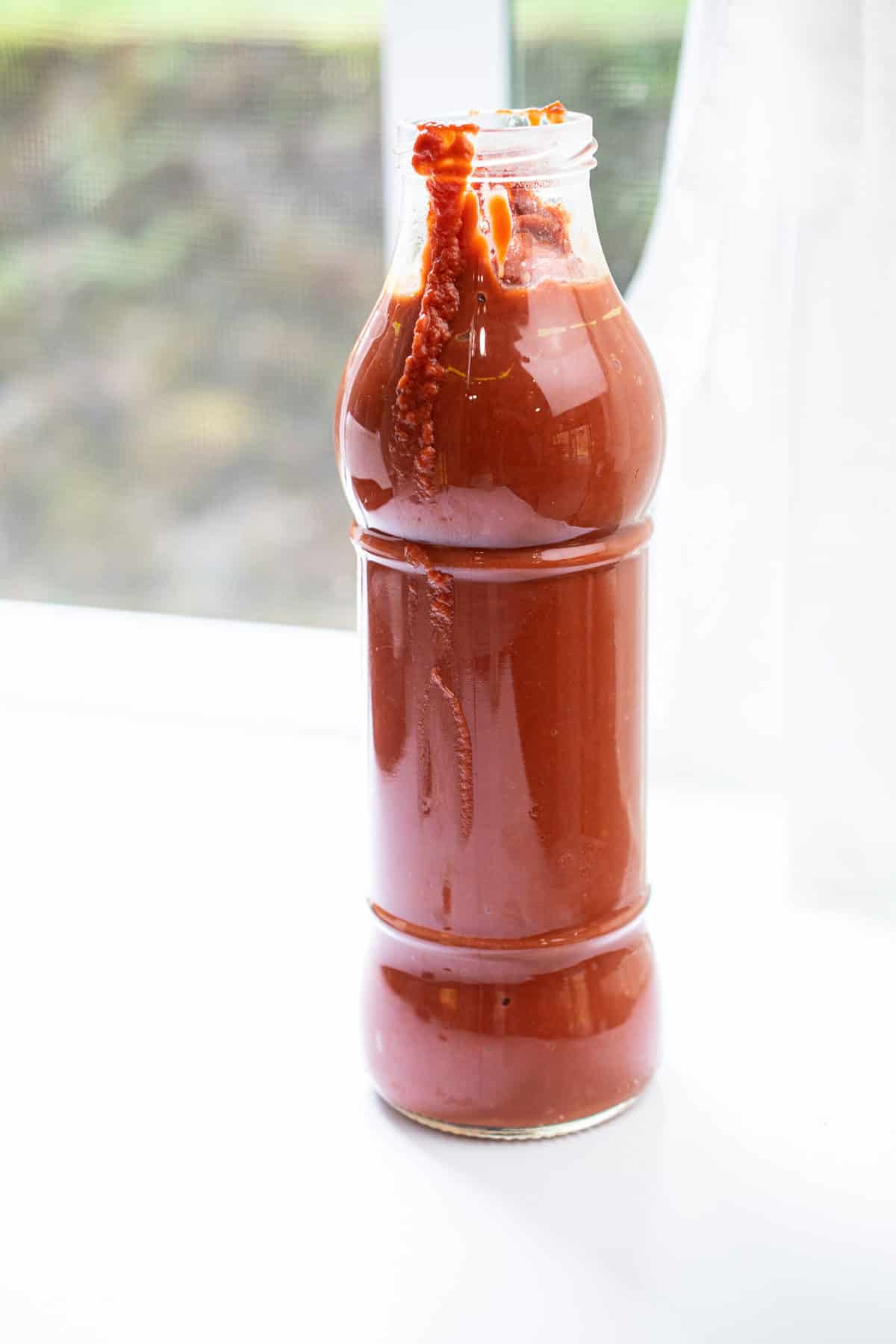 a glass marinara bottle filled with aip nomato sauce on a white counter next to a window