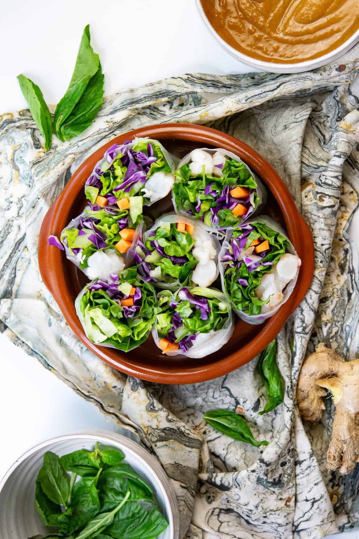 paleo spring rolls cut in half and tucked into brown ceramic bowl over a napkin with basil and ginger around them