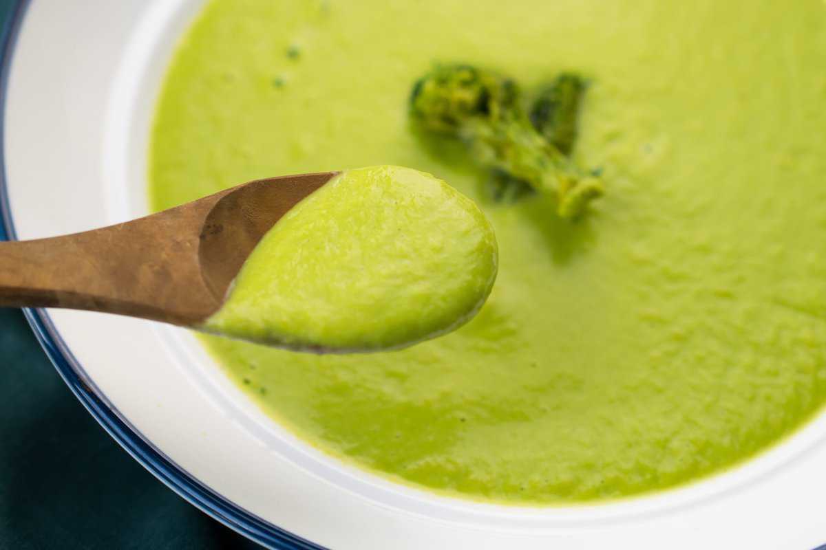 a wooden spoon scooping creamy whole30 asparagus soup from a white bowl, it's bright green and creamy 