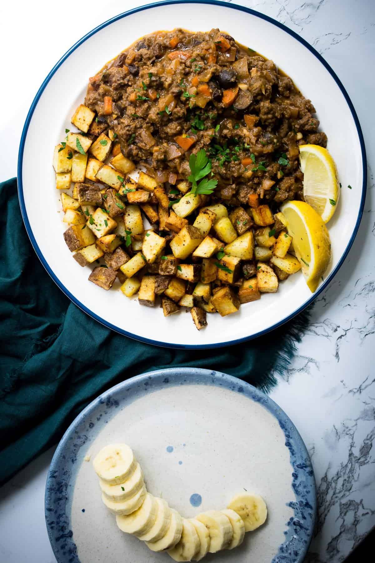 plate of cuban picadillo served with potatoes on a marble counter a plate of sliced bananas on the side 