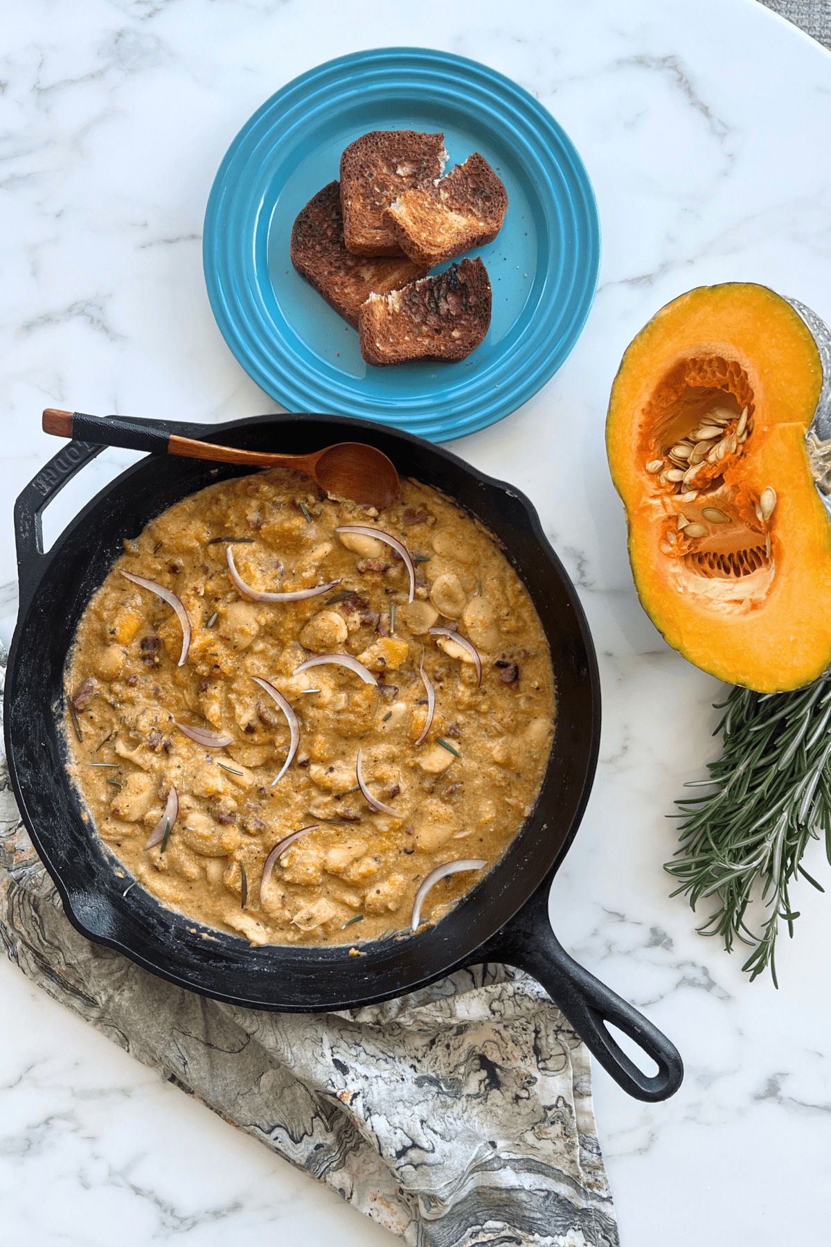 a skillet with creamy pumpkin butter beans on a white marble table next time half a pumpkin, a plate with toast and a bundle of herbs